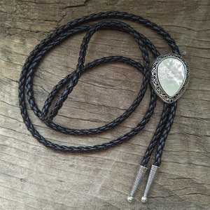 May include: A black braided bolo tie with a silver and white pearlescent pendant. The pendant has a decorative filigree design.