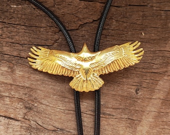 May include: A gold-coloured bolo tie featuring a detailed eagle with outstretched wings. The eagle pendant is suspended from a black cord, set against a rustic wooden background. The eagle's wingspan is approximately 7.6 cm.