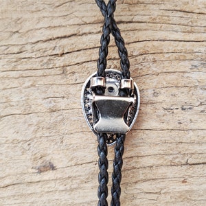 May include: A black braided bolo tie with a silver-toned metal clasp in the shape of a heart. The bolo tie is on a wooden surface.
