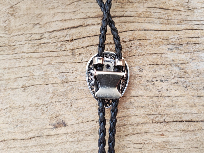 May include: A black braided bolo tie with a silver-toned metal clasp in the shape of a heart. The bolo tie is on a wooden surface.