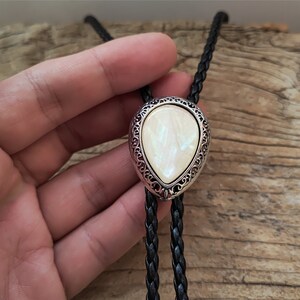 May include: A black leather bolo tie with a silver-toned pendant featuring a white iridescent stone. The pendant has a decorative filigree design.