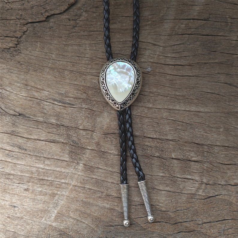 May include: A silver bolo tie with a white pearlescent stone set in a decorative frame. The tie is made of black braided leather.