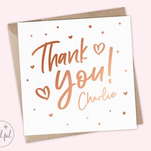 Puede incluir: Una tarjeta blanca con la frase "Thank You!" en letras de color cobre y el nombre "Charlie". Peque&ntilde;os corazones y puntos de color cobre decoran la tarjeta. Se presenta dentro de un sobre marr&oacute;n.