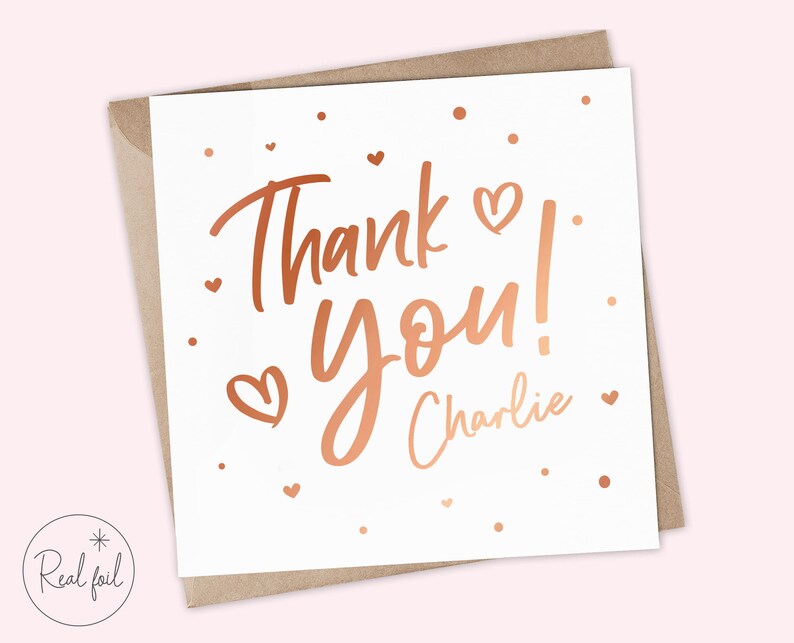 Puede incluir: Una tarjeta blanca con la frase "Thank You!" en letras de color cobre y el nombre "Charlie". Peque&ntilde;os corazones y puntos de color cobre decoran la tarjeta. Se presenta dentro de un sobre marr&oacute;n.