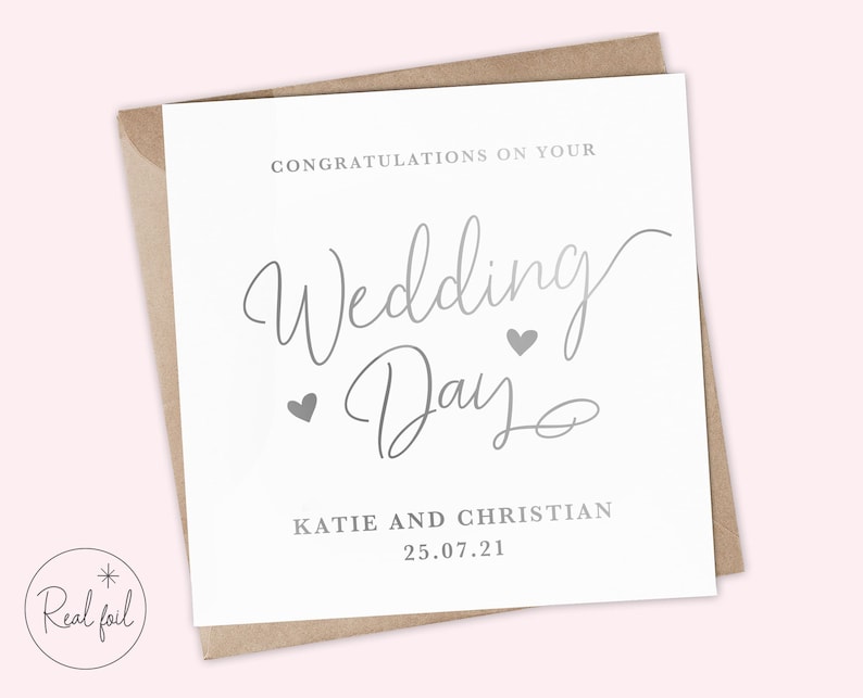 K&ouml;nnte beinhalten: Eine wei&szlig;e Hochzeitskarte mit silberner Folienbeschriftung, auf der "Herzlichen Gl&uuml;ckwunsch zu Ihrem Hochzeitstag" mit zwei kleinen Herzen steht. Die Karte enth&auml;lt auch die Namen Katie und Christian sowie das Datum 25.07.21.