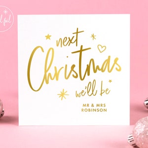May include: A white square card with gold foil text that reads "next Christmas we'll be" with the names "MR & MRS ROBINSON" below. The card is decorated with gold stars and a heart. The background is pink, with silver Christmas ornaments.