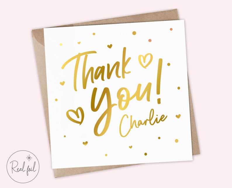 Puede incluir: Tarjeta de felicitaci&oacute;n blanca con letras doradas que dicen "Thank You!" y el nombre "Charlie". La tarjeta presenta detalles de corazones dorados y est&aacute; colocada en un sobre marr&oacute;n. Las palabras "Real foil" est&aacute;n impresas en un c&iacute;rculo en la esquina inferior izquierda.