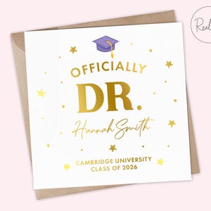 May include: A white graduation card with gold foil text reading "OFFICIALLY DR. Hannah Smith" and "CAMBRIDGE UNIVERSITY CLASS OF 2026". The card features a purple graduation cap illustration and gold star accents.