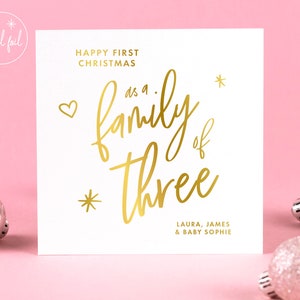 May include: A white Christmas card with gold foil lettering. The card reads "Happy First Christmas as a family of three" with the names Laura, James & Baby Sophie. Decorative heart and star accents are included.