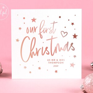 May include: A white square Christmas card with rose gold foil text that reads "our first Christmas". The card includes the names "AS MR & MRS THOMPSON" and the year "2020". The card is decorated with stars and a heart.