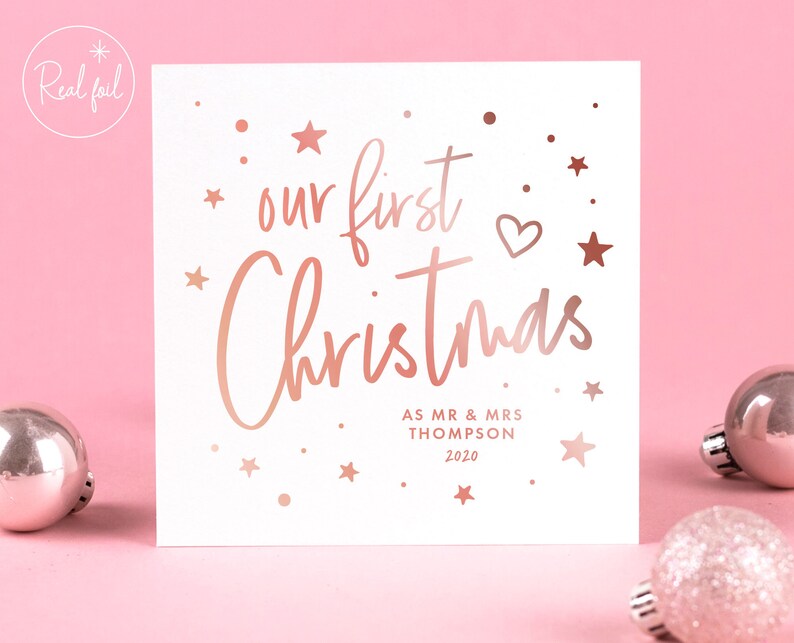 May include: A white square Christmas card with rose gold foil text that reads "our first Christmas". The card includes the names "AS MR & MRS THOMPSON" and the year "2020". The card is decorated with stars and a heart.