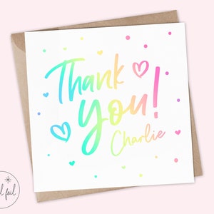 Puede incluir: Tarjeta de felicitaci&oacute;n blanca con un mensaje "Thank You!" en degradado arco&iacute;ris y el nombre "Charlie" en escritura cursiva. La tarjeta presenta peque&ntilde;os corazones y puntos de varios colores. Se encuentra dentro de un sobre marr&oacute;n.