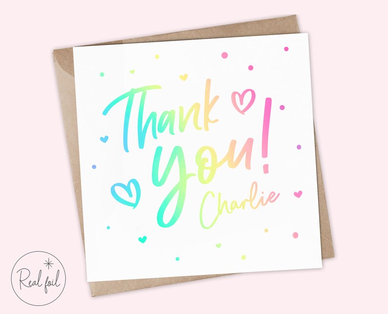 Puede incluir: Tarjeta de felicitaci&oacute;n blanca con un mensaje "Thank You!" en degradado arco&iacute;ris y el nombre "Charlie" en escritura cursiva. La tarjeta presenta peque&ntilde;os corazones y puntos de varios colores. Se encuentra dentro de un sobre marr&oacute;n.