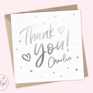 Puede incluir: Tarjeta blanca con letras plateadas que dicen "Thank you! Charlie". La tarjeta tiene peque&ntilde;os corazones y puntos plateados. Se presenta dentro de un sobre marr&oacute;n.