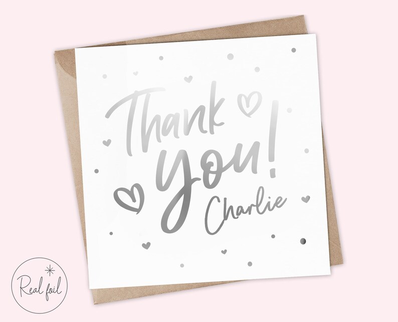 Puede incluir: Tarjeta blanca con letras plateadas que dicen "Thank you! Charlie". La tarjeta tiene peque&ntilde;os corazones y puntos plateados. Se presenta dentro de un sobre marr&oacute;n.