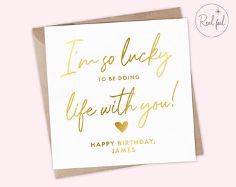May include: A white greeting card with gold foil lettering. The text reads "I'm so lucky to be doing life with you!" and "Happy Birthday, James." A gold heart is also featured. The card is inside a brown envelope.