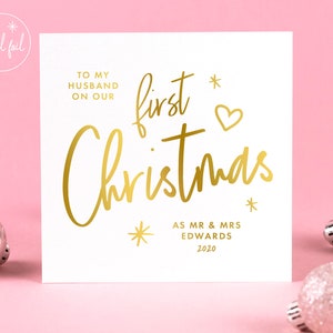 May include: A white square Christmas card with gold foil lettering. The text reads "To my husband on our first Christmas" with the names "Mr & Mrs Edwards" and the year "2020" below. The card has a pink background with silver ornaments.