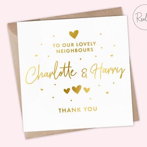 Puede incluir: Una tarjeta de felicitación blanca con texto dorado y detalles de corazón. La tarjeta dice "TO OUR LOVELY NEIGHBOURS Charlotte & Harry THANK YOU". La tarjeta está dentro de un sobre marrón y tiene un logo "Real foil".