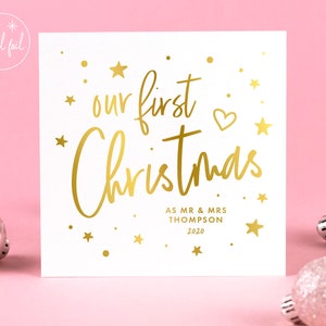 May include: A white square card with gold foil text that reads "our first Christmas" with a heart and star accents. Below the text is "AS MR & MRS THOMPSON 2020". The card is on a pink background with silver ornaments.