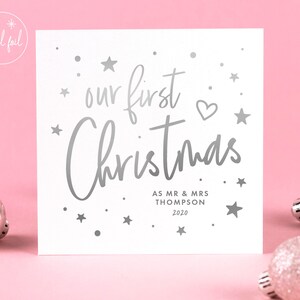 May include: A white square Christmas card with silver foil text that reads "our first Christmas" with the names "AS MR & MRS THOMPSON 2020". The card is decorated with silver stars and a heart. The background is pink.