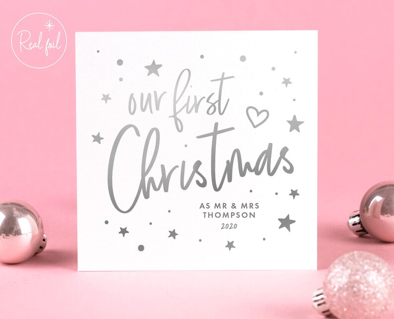 May include: A white square Christmas card with silver foil text that reads "our first Christmas" with the names "AS MR & MRS THOMPSON 2020". The card is decorated with silver stars and a heart. The background is pink.