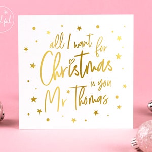 May include: A white Christmas card with gold foil lettering that reads "all I want for Christmas is you Mr Thomas." Gold stars and a heart accent the text. The card is on a pink background with silver ornaments.