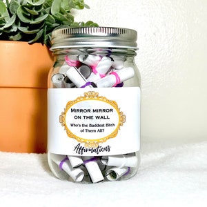 May include: A glass jar filled with rolled up pieces of paper with purple and pink rubber bands. The jar has a white label with the text "Mirror Mirror On The Wall Who's The Baddest B*tch Of Them All?" and the word "Affirmations" below the text.