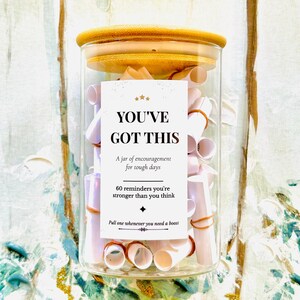 May include: A clear glass jar with a wooden lid filled with small white paper scrolls secured by orange rubber bands. A white label on the front reads: YOU'VE GOT THIS, A jar of encouragement for tough days, 60 reminders you're stronger than you think.