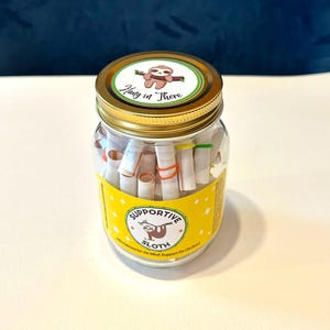 May include: A clear glass jar with a gold lid, filled with rolled-up paper scrolls. The jar has a yellow label with a sloth illustration and the words "Supportive Sloth." The lid features a sloth and the phrase "Hang in There."