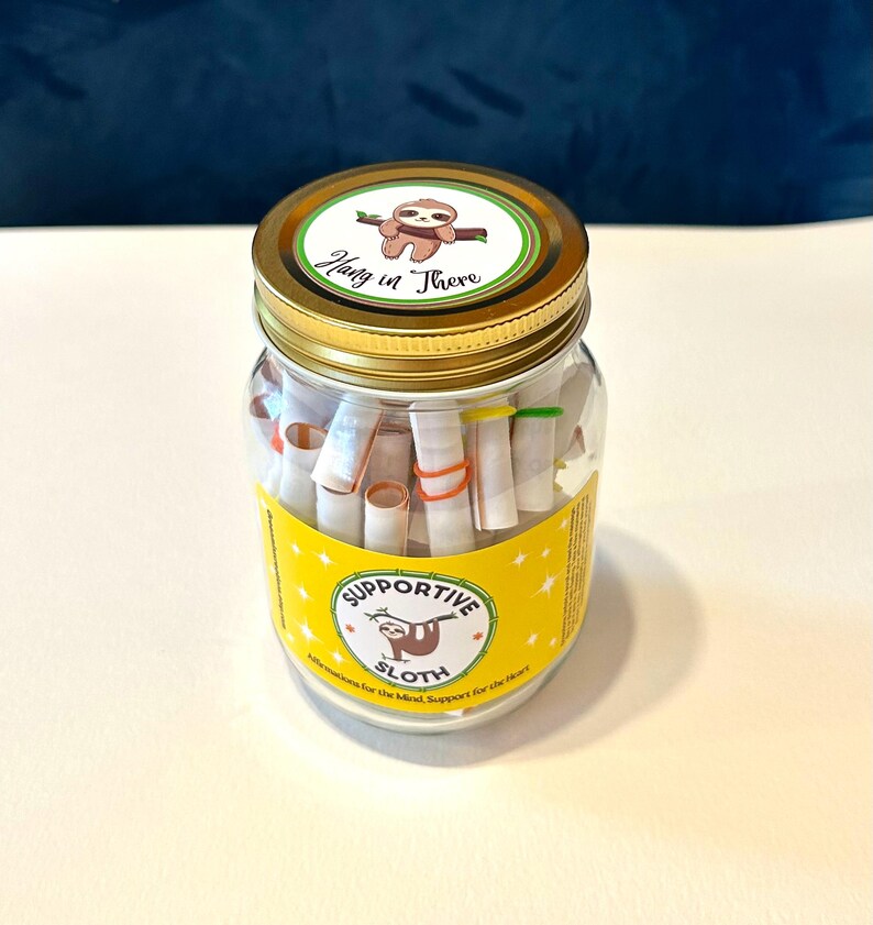 May include: A clear glass jar with a gold lid, filled with rolled-up paper scrolls. The jar has a yellow label with a sloth illustration and the words "Supportive Sloth." The lid features a sloth and the phrase "Hang in There."