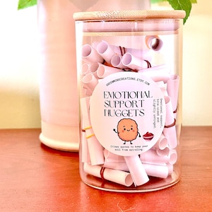 May include: A clear glass jar with a bamboo lid filled with small white paper scrolls secured by thin rubber bands. A circular label on the front reads: GREENMINXCREATIONS.ETSY.COM, EMOTIONAL SUPPORT NUGGETS, Emotional support, bite-sized and slightly unhinged, Crispy quotes to keep your soul from spiraling, with a cartoon nugget illustration.
