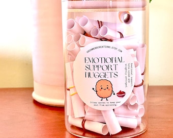 May include: A clear glass jar with a bamboo lid filled with small white paper scrolls secured by thin rubber bands. A circular label on the front reads: GREENMINXCREATIONS.ETSY.COM, EMOTIONAL SUPPORT NUGGETS, Emotional support, bite-sized and slightly unhinged, Crispy quotes to keep your soul from spiraling, with a cartoon nugget illustration.
