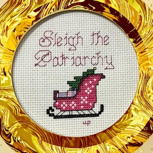 Op de afbeelding: Een witte kruissteek met een roze en witte slee met een groene boom erop. De tekst "Sleigh the Patriarchy" is in paars boven de slee geborduurd.
