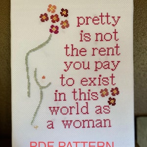 May include: Cross-stitch artwork on a white canvas. The design features a female form with floral accents and the text "pretty is not the rent you pay to exist in this world as a woman." The words "PDF PATTERN" are at the bottom.