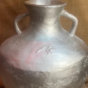 Puede incluir: Un jarrón plateado hecho a mano, con dos asas y tres patas. El jarrón tiene un cuerpo redondeado y una boca ancha. Un pequeño emblema es visible en la parte delantera del jarrón. El jarrón está siendo sostenido por una persona.