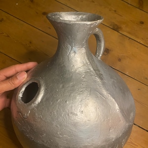Puede incluir: Una jarra de cerámica pintada de plata con cuerpo redondo y una pequeña asa. La jarra tiene un agujero en el lateral.