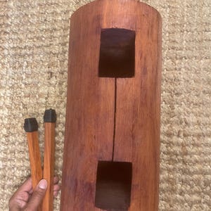 Op de afbeelding: Een cilindrische houten spleet-trommel met een roodbruine afwerking, voorzien van twee rechthoekige uitsparingen aan de voorzijde en een handvat aan de bovenkant. Twee bijpassende houten stokken met donkere doppen liggen naast het instrument.