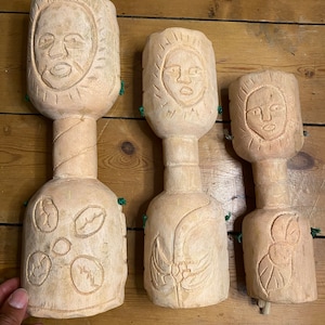 May include: Set of three wooden carved figures with human faces and floral designs. The figures are different sizes and have a rustic, handmade look.