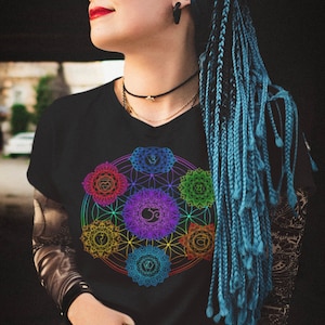 May include: A woman with blue braids wears a black t-shirt with a colorful chakra symbol design. The symbol is a circle with seven smaller circles inside, each a different color of the rainbow.