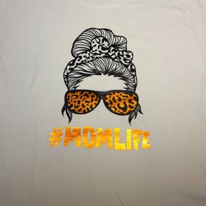 Peut inclure: T-shirt blanc avec un motif noir et orange d'une femme avec un chignon d&eacute;sordonn&eacute;, des lunettes de soleil et le texte "#MOMLIFE" en or.