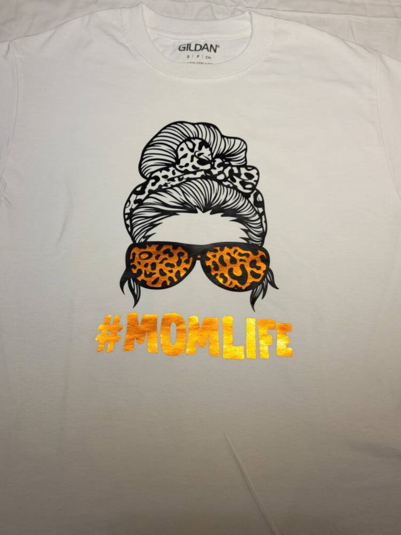 Peut inclure: T-shirt blanc avec un motif noir et orange d'une femme avec un chignon d&eacute;sordonn&eacute;, des lunettes de soleil et le texte "#MOMLIFE" en or.