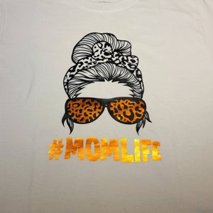 Peut inclure: T-shirt blanc avec un motif noir et orange d'une femme avec un chignon d&eacute;sordonn&eacute;, des lunettes de soleil et le texte "#MOMLIFE" en or.
