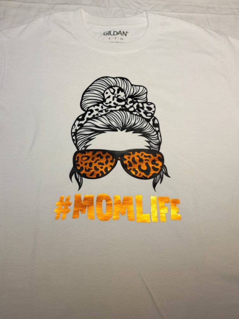Peut inclure: T-shirt blanc avec un motif noir et orange d'une femme avec un chignon d&eacute;sordonn&eacute;, des lunettes de soleil et le texte "#MOMLIFE" en or.