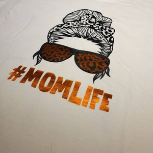 Peut inclure: T-shirt blanc avec une illustration en noir et blanc d'une femme avec un chignon et des lunettes de soleil. Le texte "#MOMLIFE" est imprim&eacute; en orange sur le t-shirt.