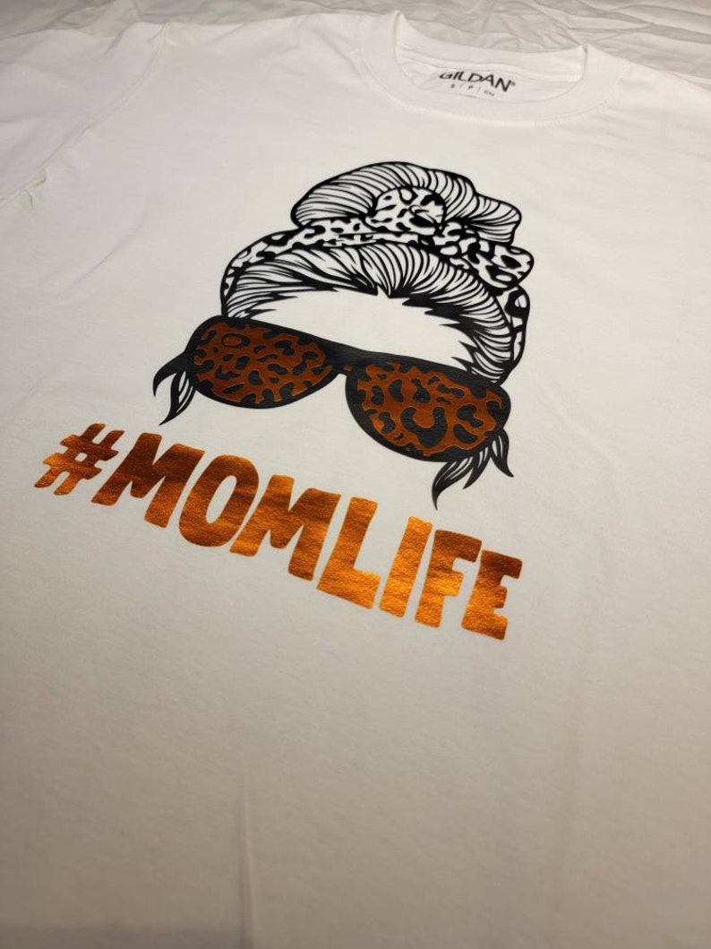 Peut inclure: T-shirt blanc avec une illustration en noir et blanc d'une femme avec un chignon et des lunettes de soleil. Le texte "#MOMLIFE" est imprim&eacute; en orange sur le t-shirt.