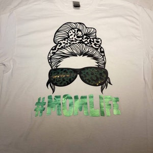 Peut inclure: T-shirt blanc avec un motif noir et blanc d'une femme avec un chignon d&eacute;sordonn&eacute;, des lunettes de soleil et un bandeau &agrave; imprim&eacute; l&eacute;opard. Le texte "#MOMLIFE" est imprim&eacute; en vert sous le motif.