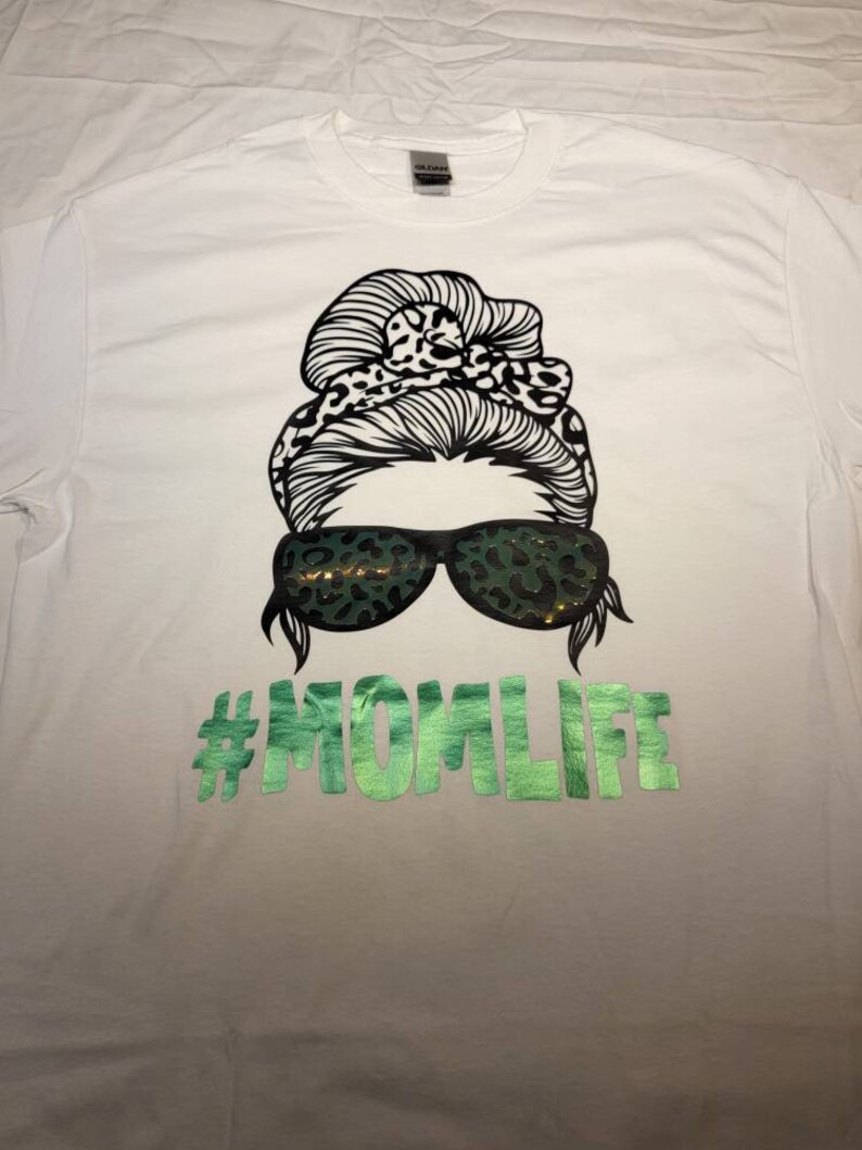 Peut inclure: T-shirt blanc avec un motif noir et blanc d'une femme avec un chignon d&eacute;sordonn&eacute;, des lunettes de soleil et un bandeau &agrave; imprim&eacute; l&eacute;opard. Le texte "#MOMLIFE" est imprim&eacute; en vert sous le motif.