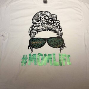 Peut inclure: T-shirt blanc avec un graphique noir et blanc d'une femme avec un chignon d&eacute;sordonn&eacute; et des lunettes de soleil. Les lunettes de soleil et le chignon ont un motif l&eacute;opard. Le texte "#MOMLIFE" est imprim&eacute; en vert sous le graphique.
