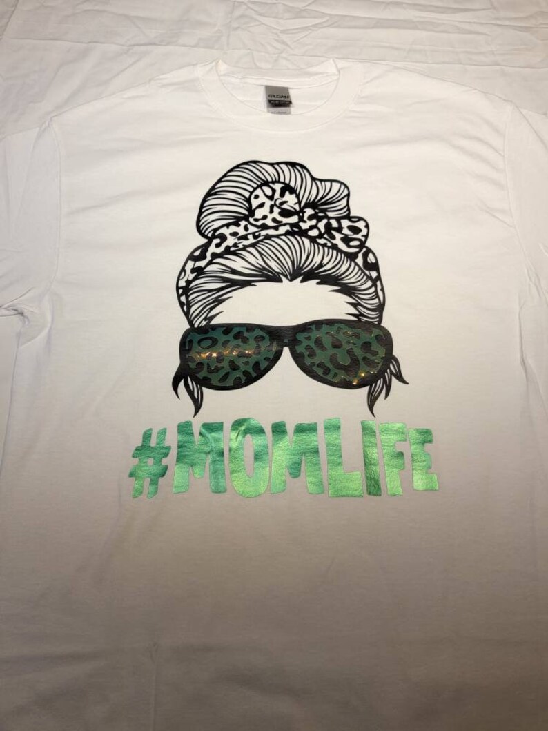 Peut inclure: T-shirt blanc avec un graphique noir et blanc d'une femme avec un chignon d&eacute;sordonn&eacute; et des lunettes de soleil. Les lunettes de soleil et le chignon ont un motif l&eacute;opard. Le texte "#MOMLIFE" est imprim&eacute; en vert sous le graphique.
