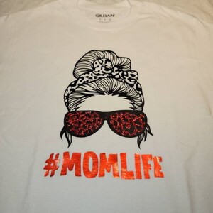 Peut inclure: T-shirt blanc avec un motif noir d'une femme avec un chignon d&eacute;sordonn&eacute;, des lunettes de soleil et le texte "#MOMLIFE" en paillettes rouges.